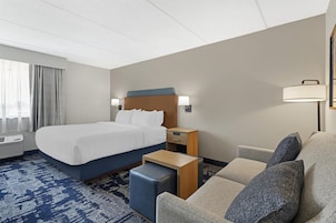 Una habitación de hotel con cama, sofá, mesita de noche, lámpara y ventana con cortinas.