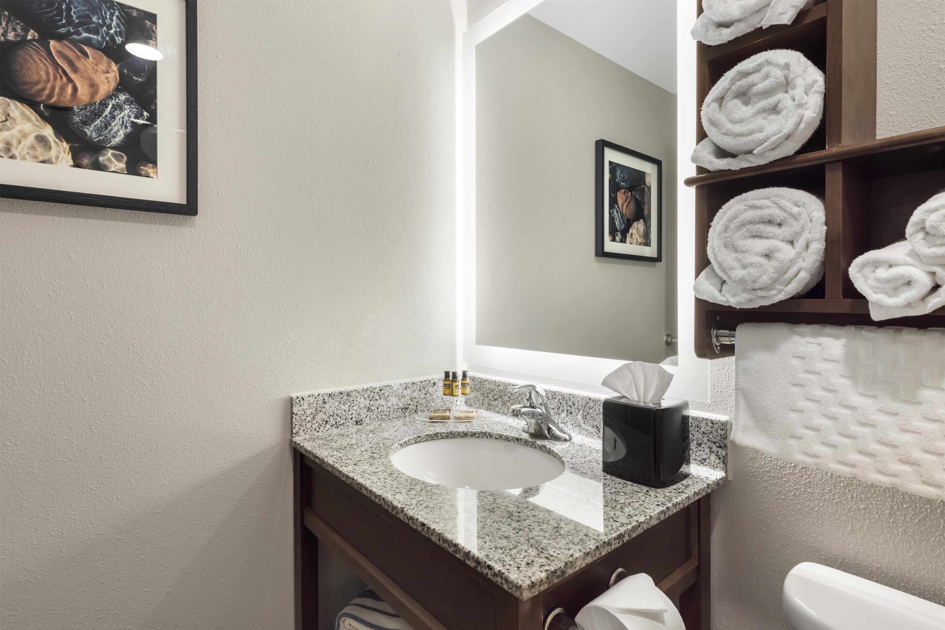 Baño con lavamanos de encimera de granito, un espejo, toallero con toallas e imagen enmarcada en la pared.