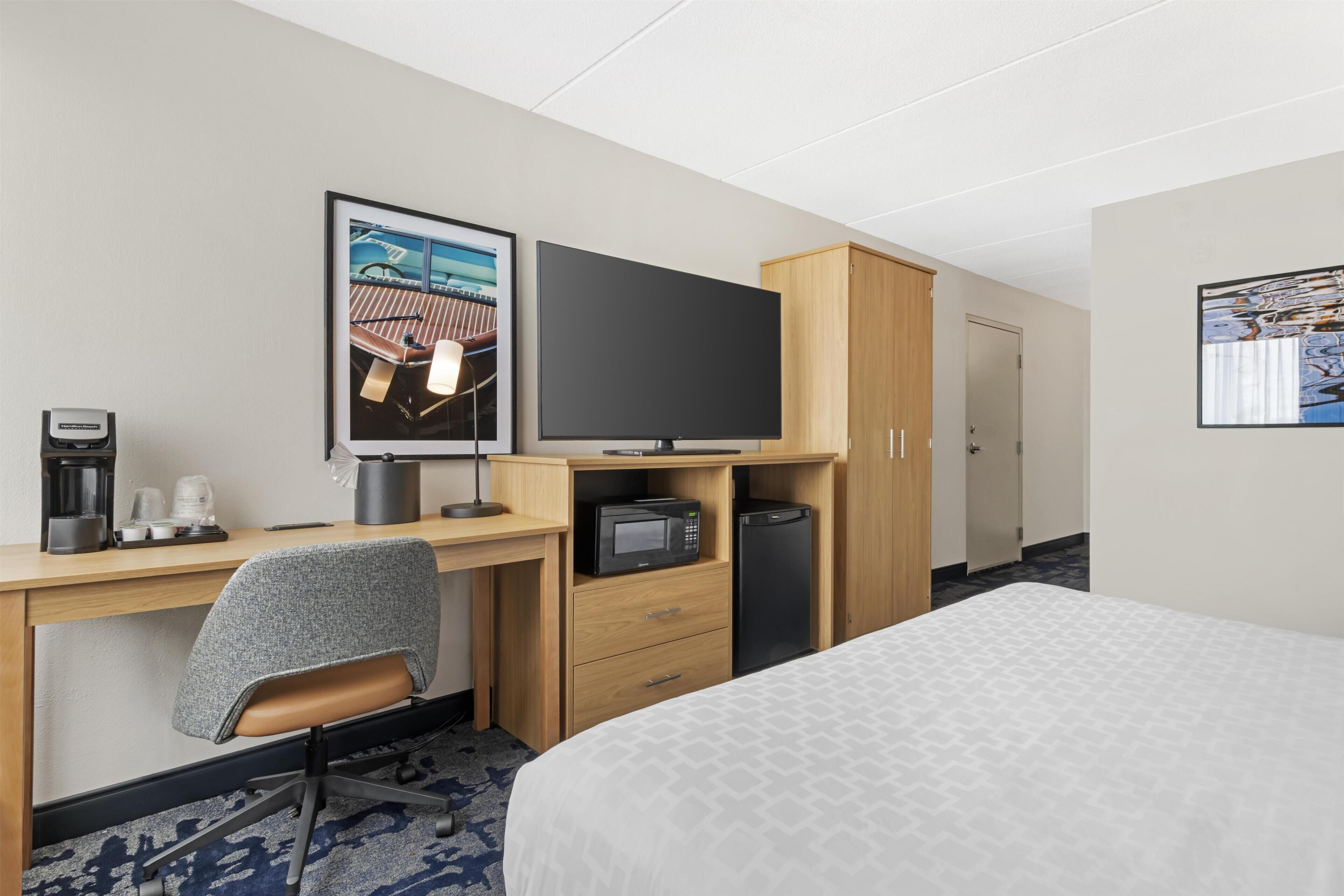 Habitación de hotel con una cama, un escritorio con silla, televisor, microondas, cafetera y un armario.