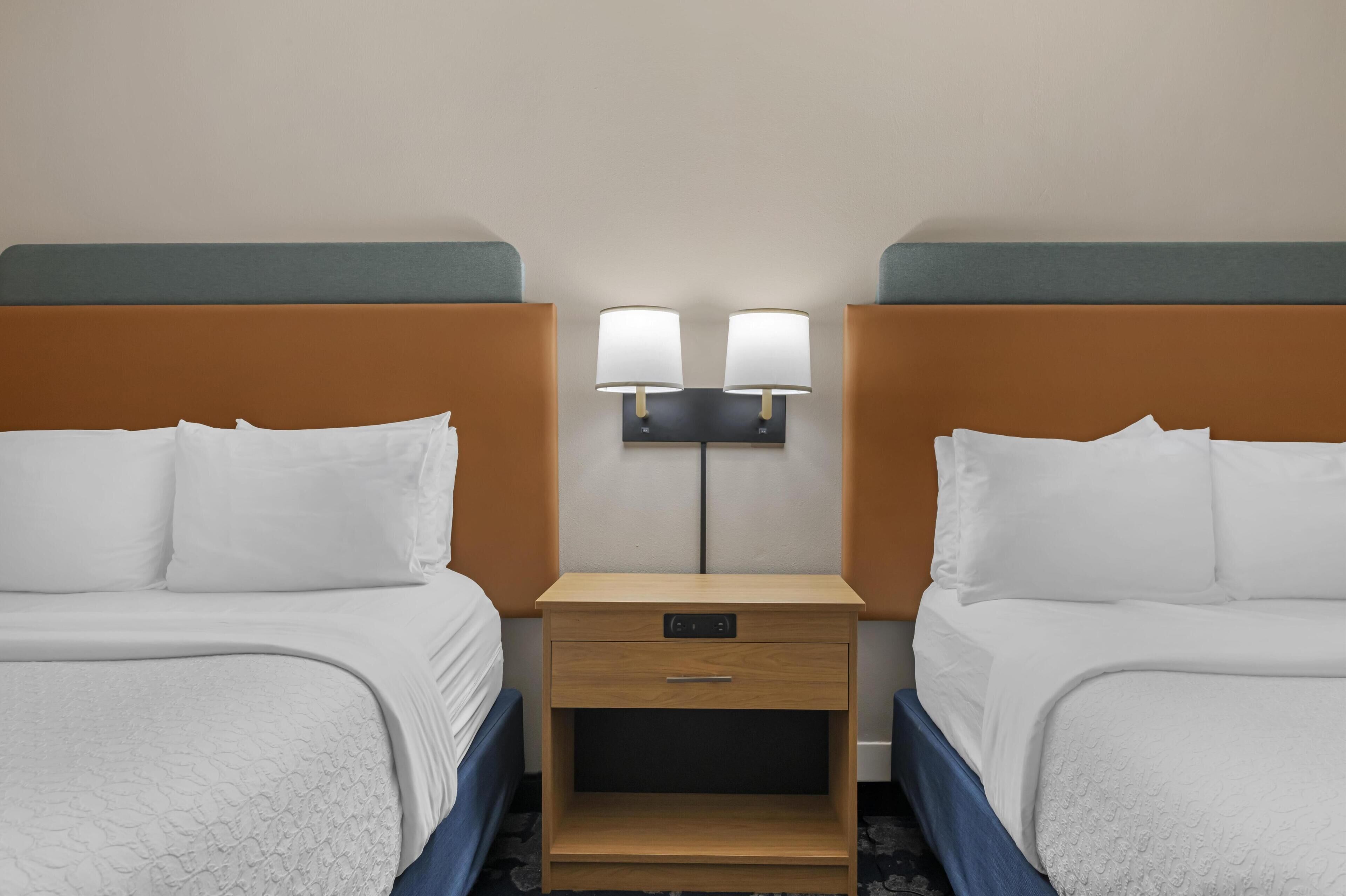 A hotel room with two beds, a nightstand with a lamp, and a small drawer.