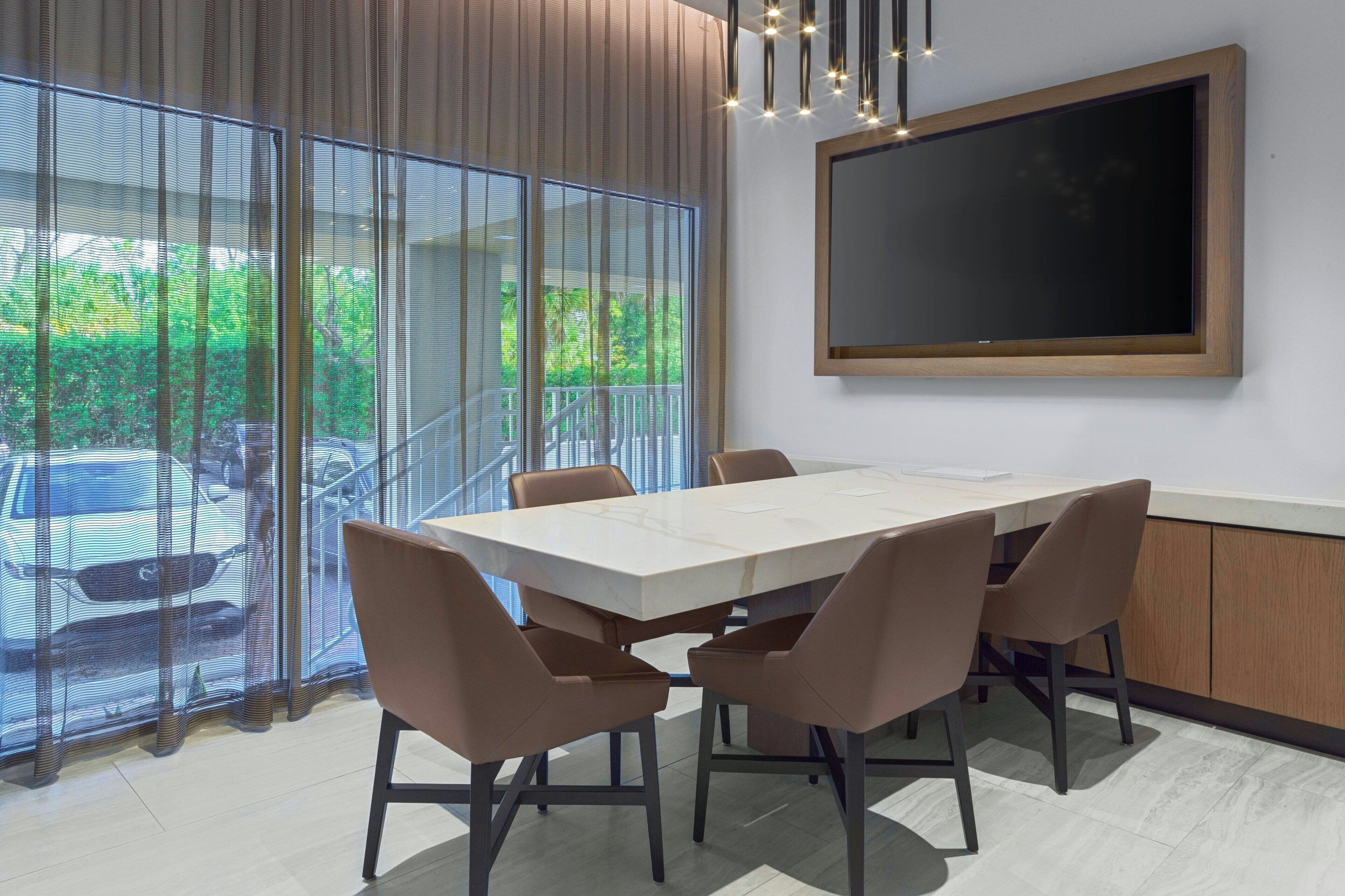 A modern conference room with a large table, six chairs, a large window with a view of greenery and parked cars, and a mounted screen on the wall.