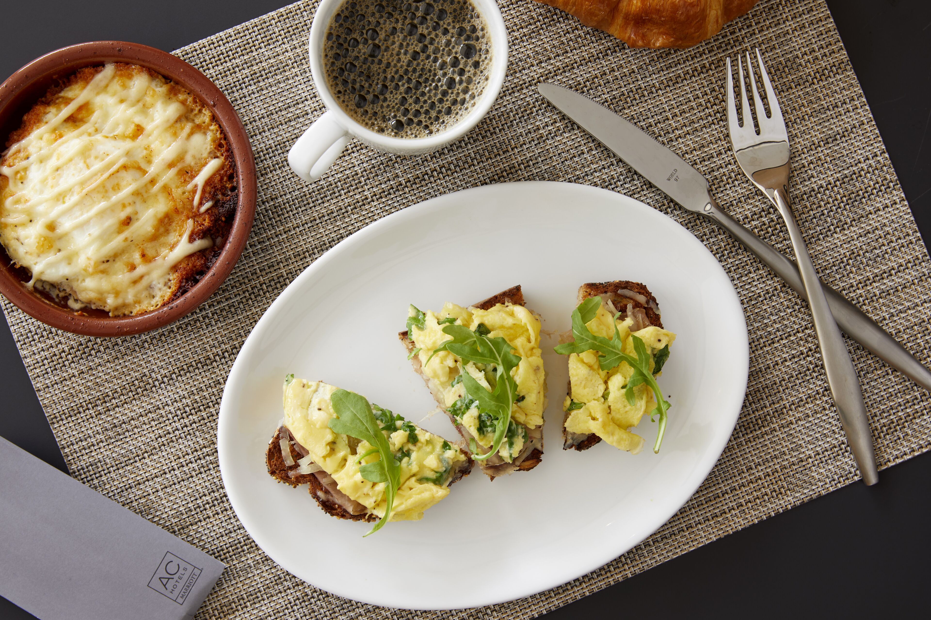 A plate with scrambled eggs and greens, a bowl of baked dish, a croissant, and breakfast utensils on a table.