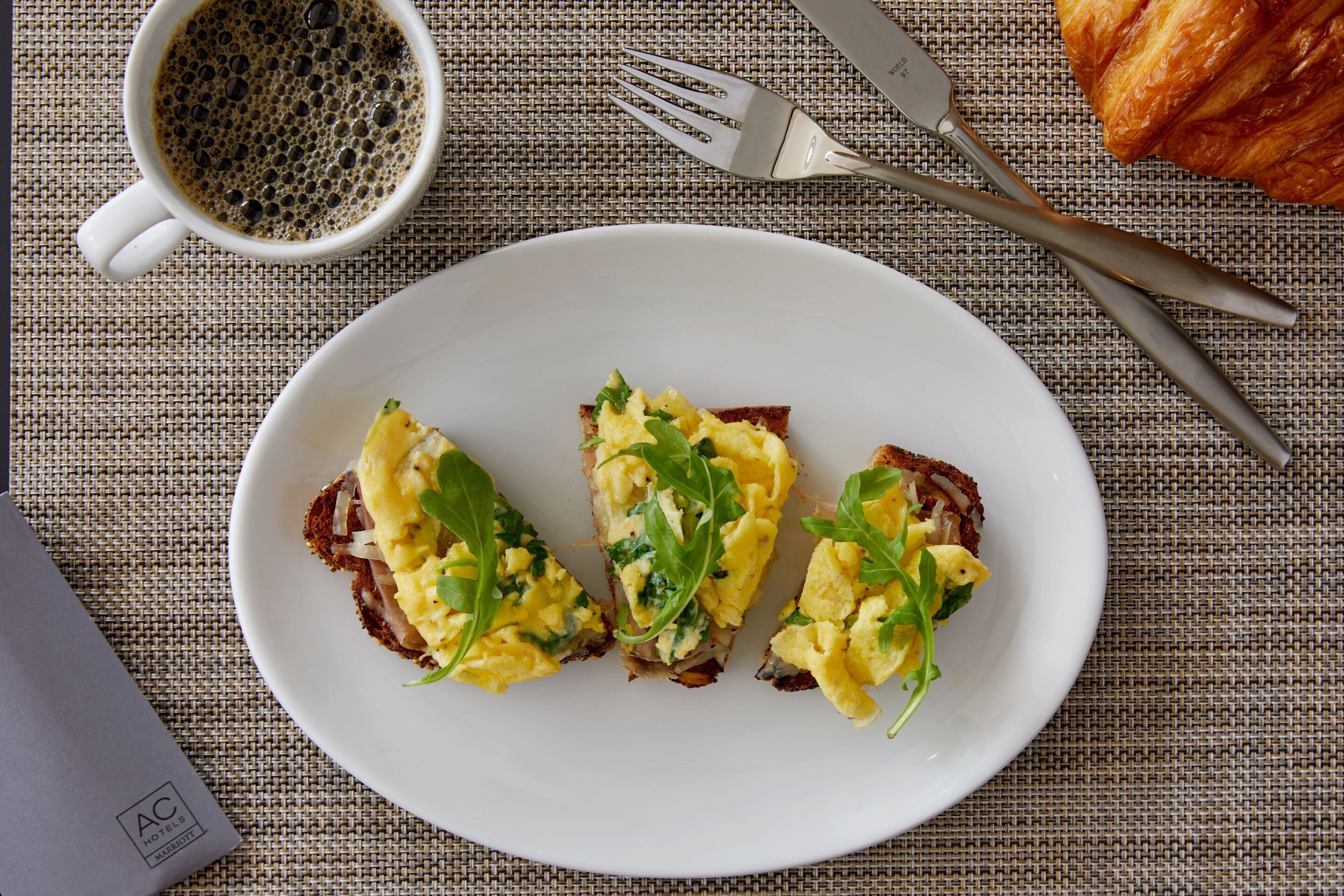 A breakfast plate with scrambled eggs and greens, a croissant, and a cup of coffee on a textured tablecloth.
