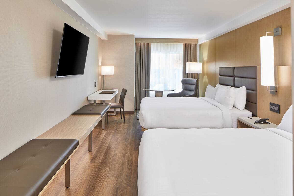 A hotel room with two beds, a desk, a chair, a TV mounted on the wall, and a window with curtains.