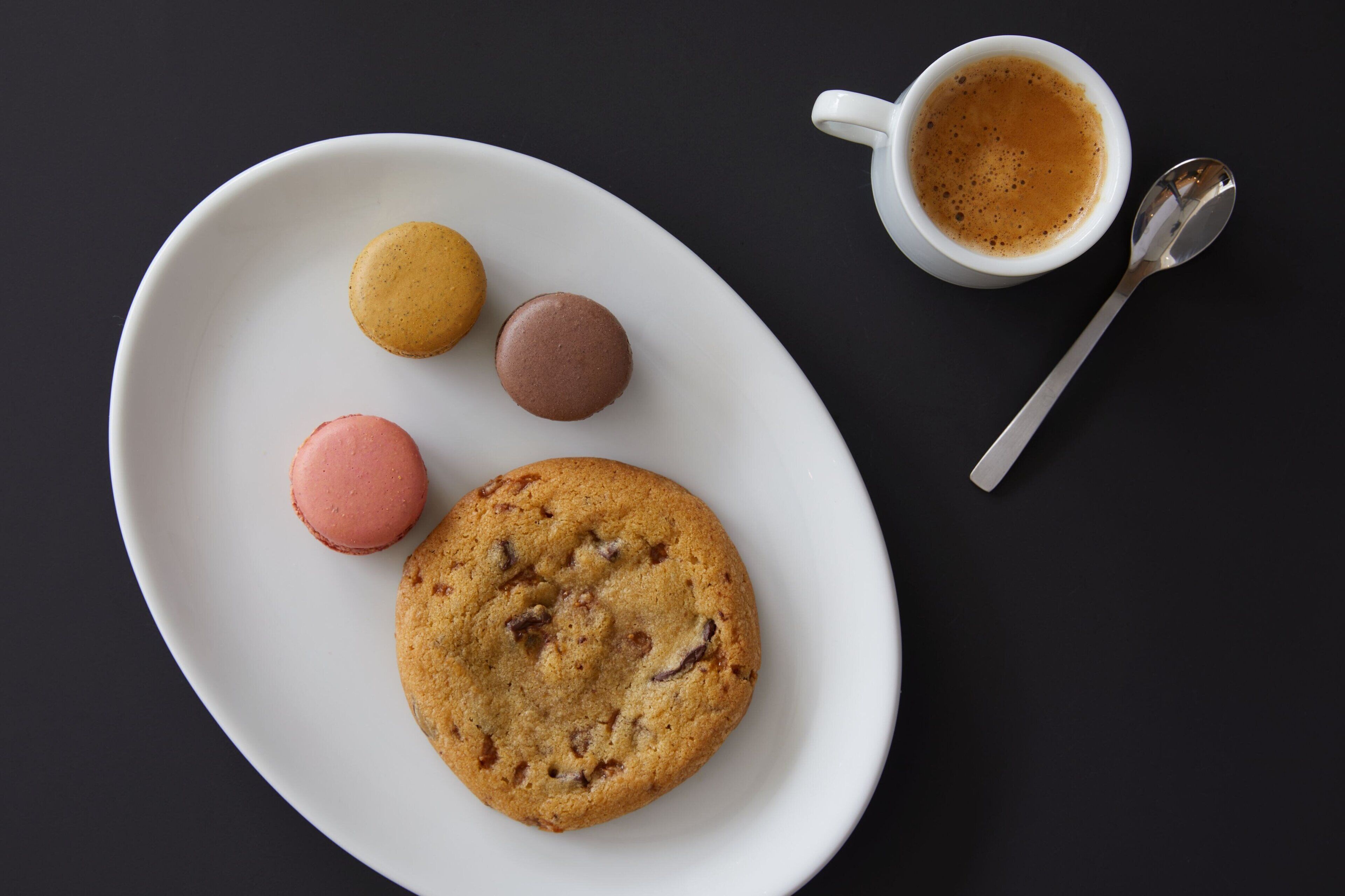 Un plato blanco con una galleta y tres macarons, una taza de café y una cuchara sobre una superficie oscura.