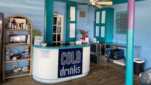 Un comptoir sur lequel est posée une enseigne indiquant « BOISSONS FRAÎCHES », dans une pièce aux murs de couleur turquoise, avec des poutres en bois et un ventilateur de plafond.