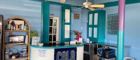 Un comptoir sur lequel est posée une enseigne indiquant « BOISSONS FRAÎCHES », dans une pièce aux murs de couleur turquoise, avec des poutres en bois et un ventilateur de plafond.