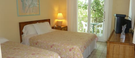 A hotel room with two beds, a nightstand with a lamp, a television, and a balcony with a view of greenery.