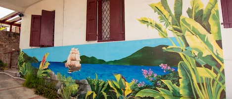 Un edificio con un mural de un barco en un mar azul, rodeado de plantas tropicales.