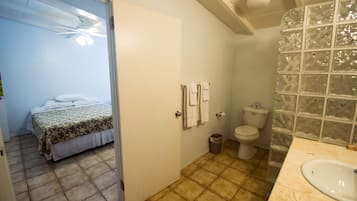 Une salle de bain équipée de toilettes, d’un lavabo et d’un espace douche. Une chambre adjacente contient un lit.