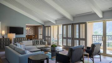 Un salon moderne avec un canapé, des fauteuils, une table basse et un téléviseur. De grandes fenêtres, équipées de stores, offrent une vue sur l’extérieur.