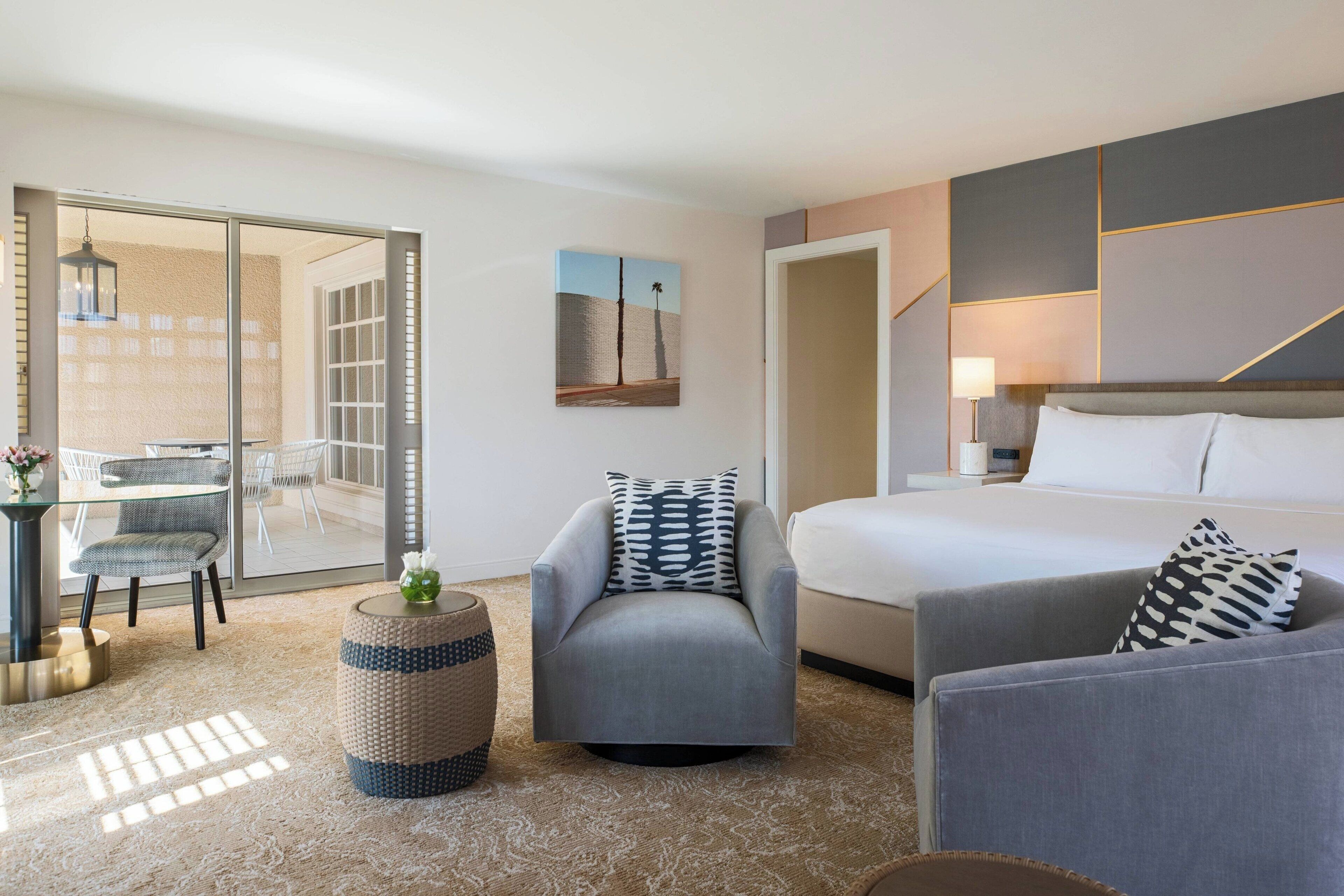 A modern hotel room with a bed, two armchairs, a small table, and a glass door leading to a balcony.