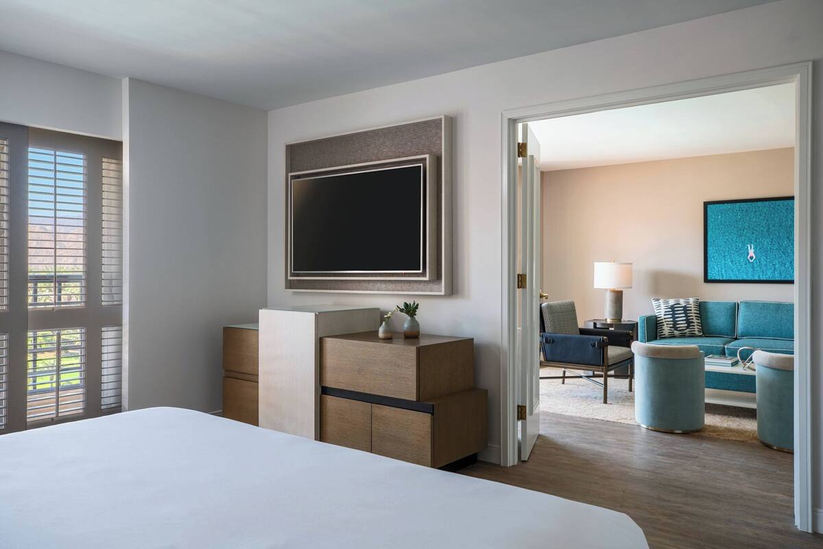 A modern hotel room with a large bed, a wooden dresser, a flat-screen TV, and a view of the living area through an open door.