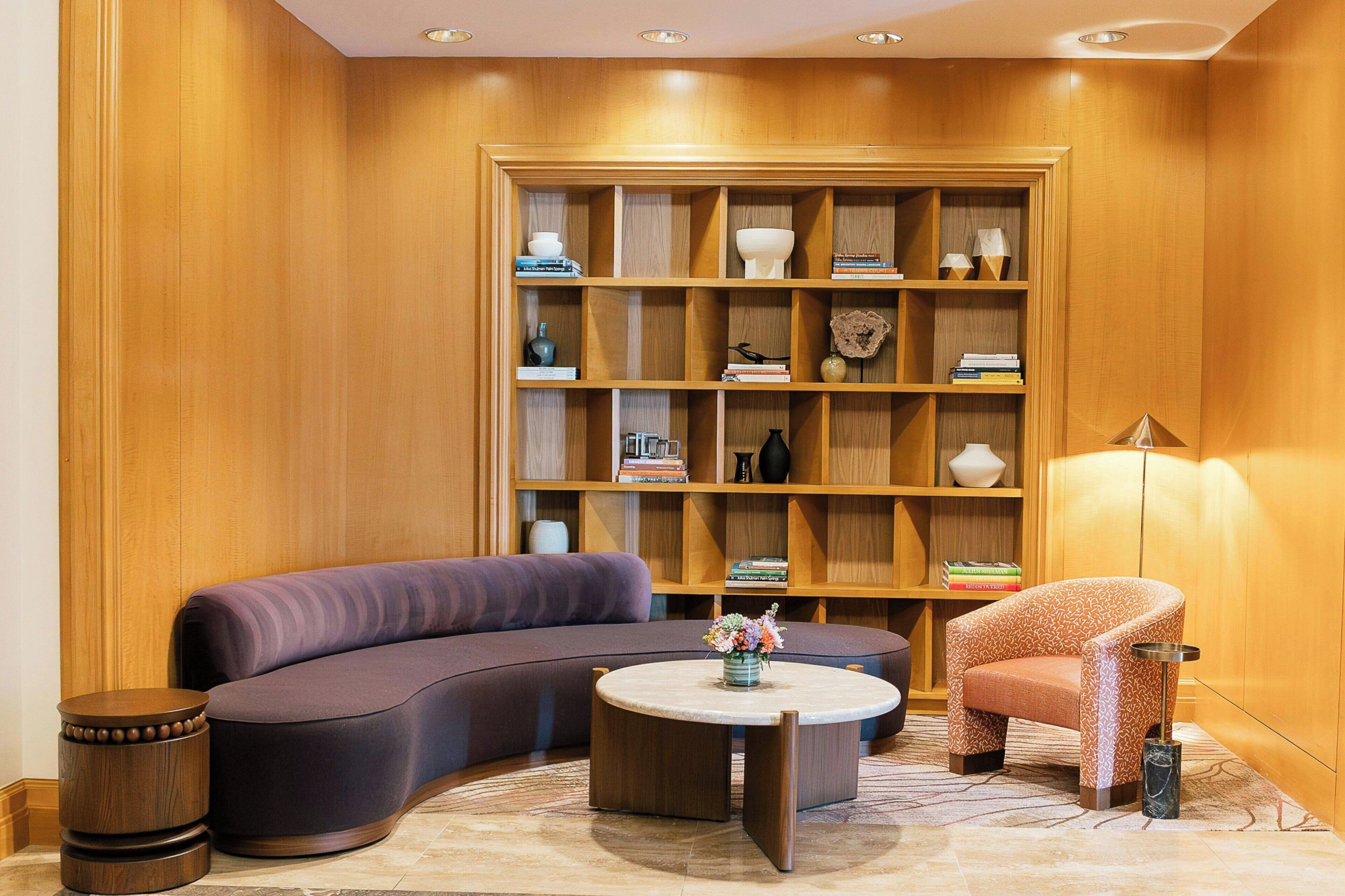 A modern living room with a curved sofa, a round coffee table, and a patterned armchair. A wooden bookshelf filled with books and decorative items is in the background.