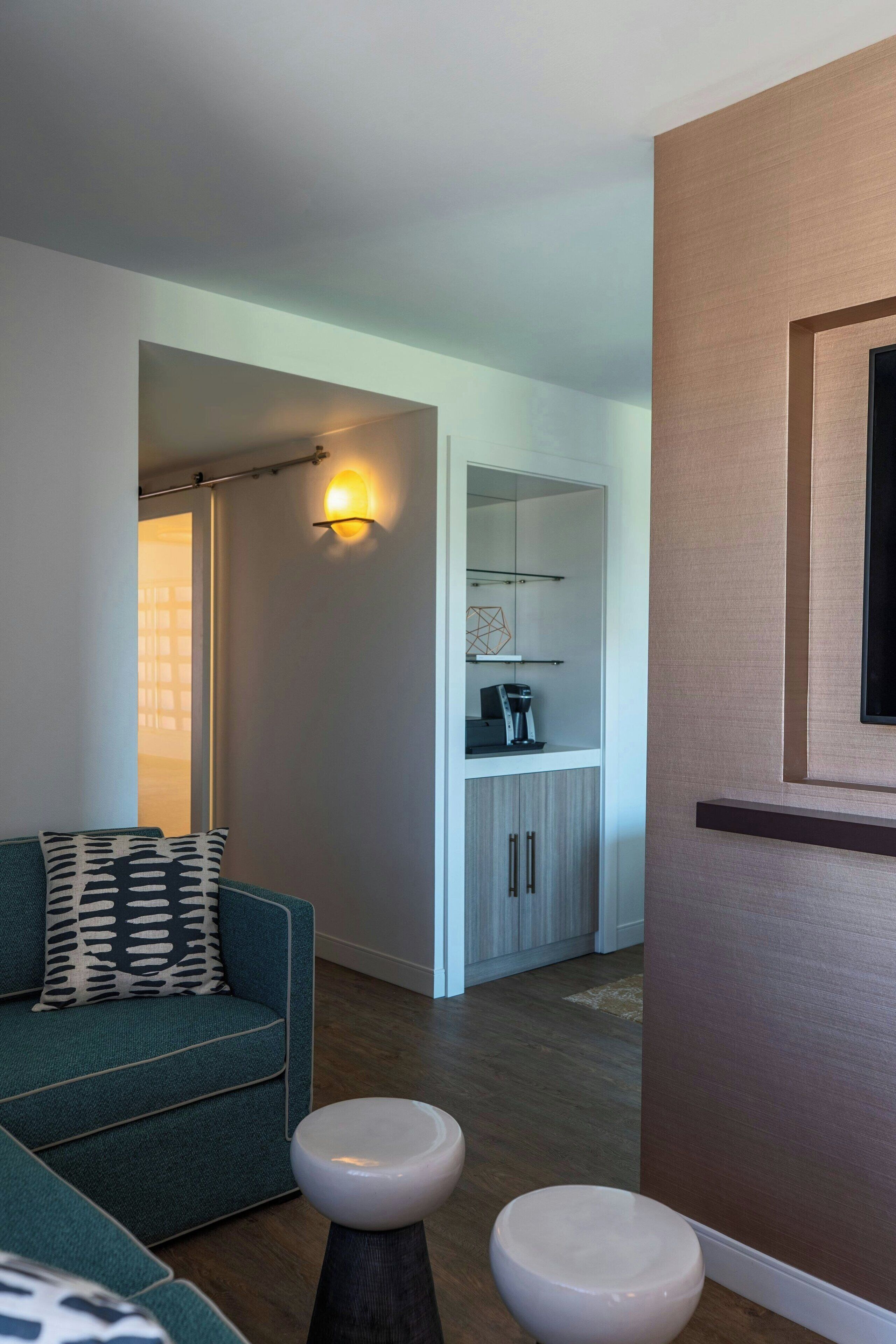 Una sala moderna con un sofá color turquesa, dos taburetes blancos, una zona de cocina con armarios y encimera, y un televisor montado en la pared.