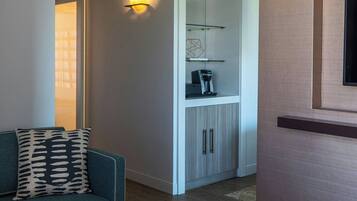 Una sala moderna con un sofá color turquesa, dos taburetes blancos, una zona de cocina con armarios y encimera, y un televisor montado en la pared.