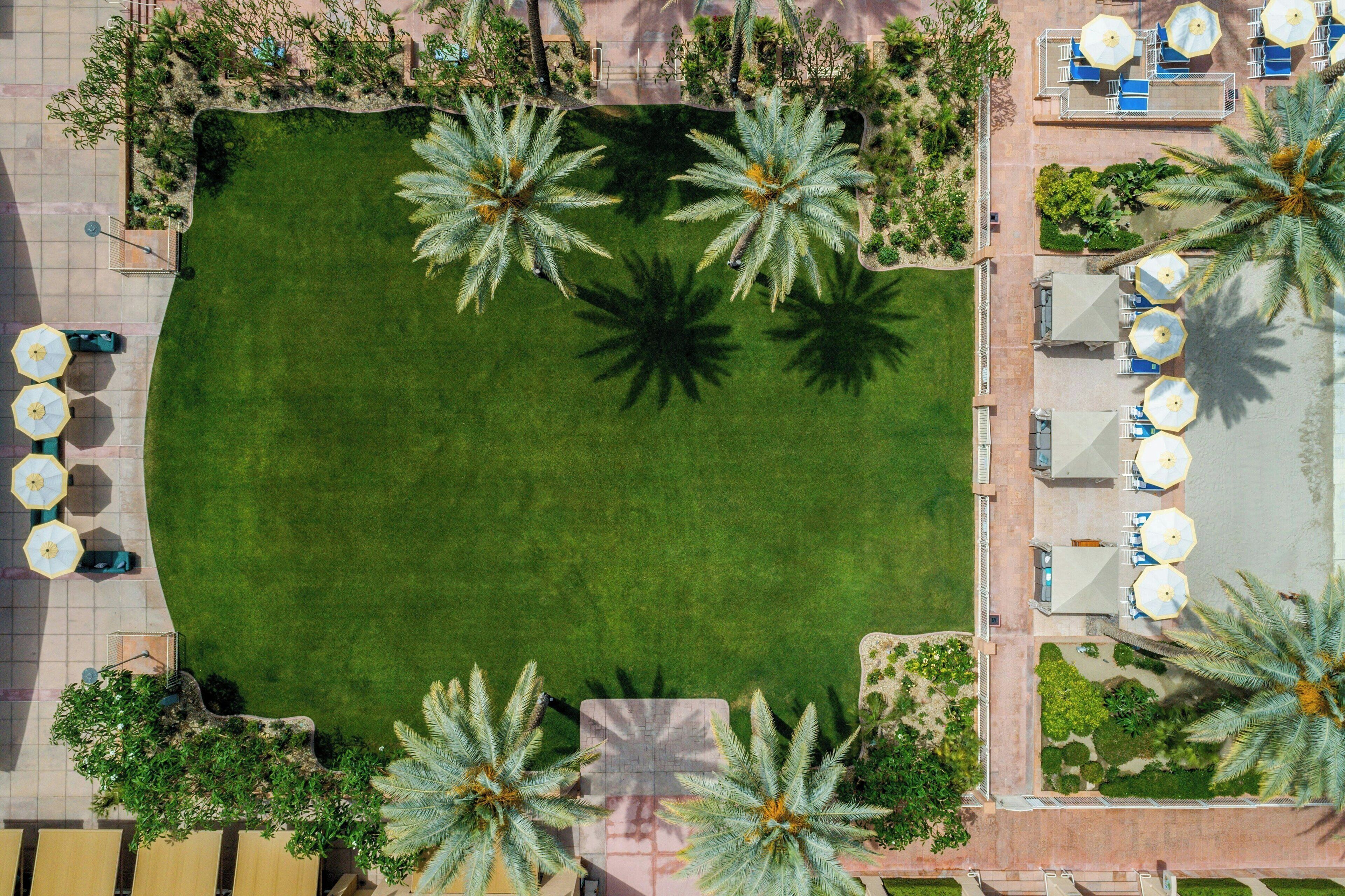 Vue aérienne d’un complexe hôtelier doté d’un espace central verdoyant, entouré de palmiers, de coins salon extérieurs avec des parasols et d’une zone piscine.