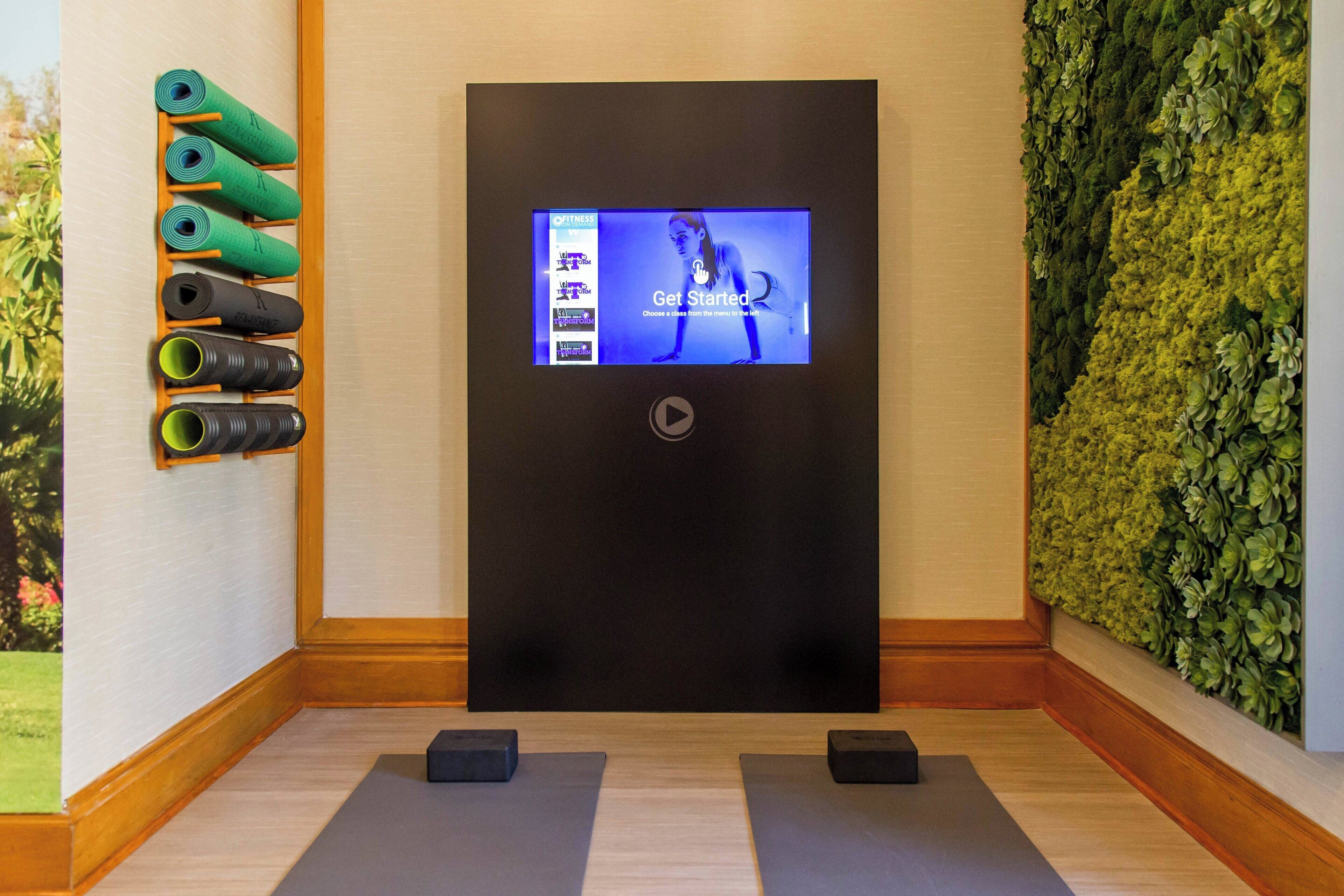 Una habitación con una pantalla digital que muestra una clase de yoga, un soporte para colchonetas de yoga en la pared, y una pared verde con follaje.