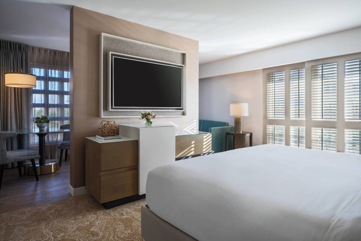 A hotel room with a large bed, a flat-screen TV mounted on the wall, a desk with a lamp, and a view of the city through large windows.