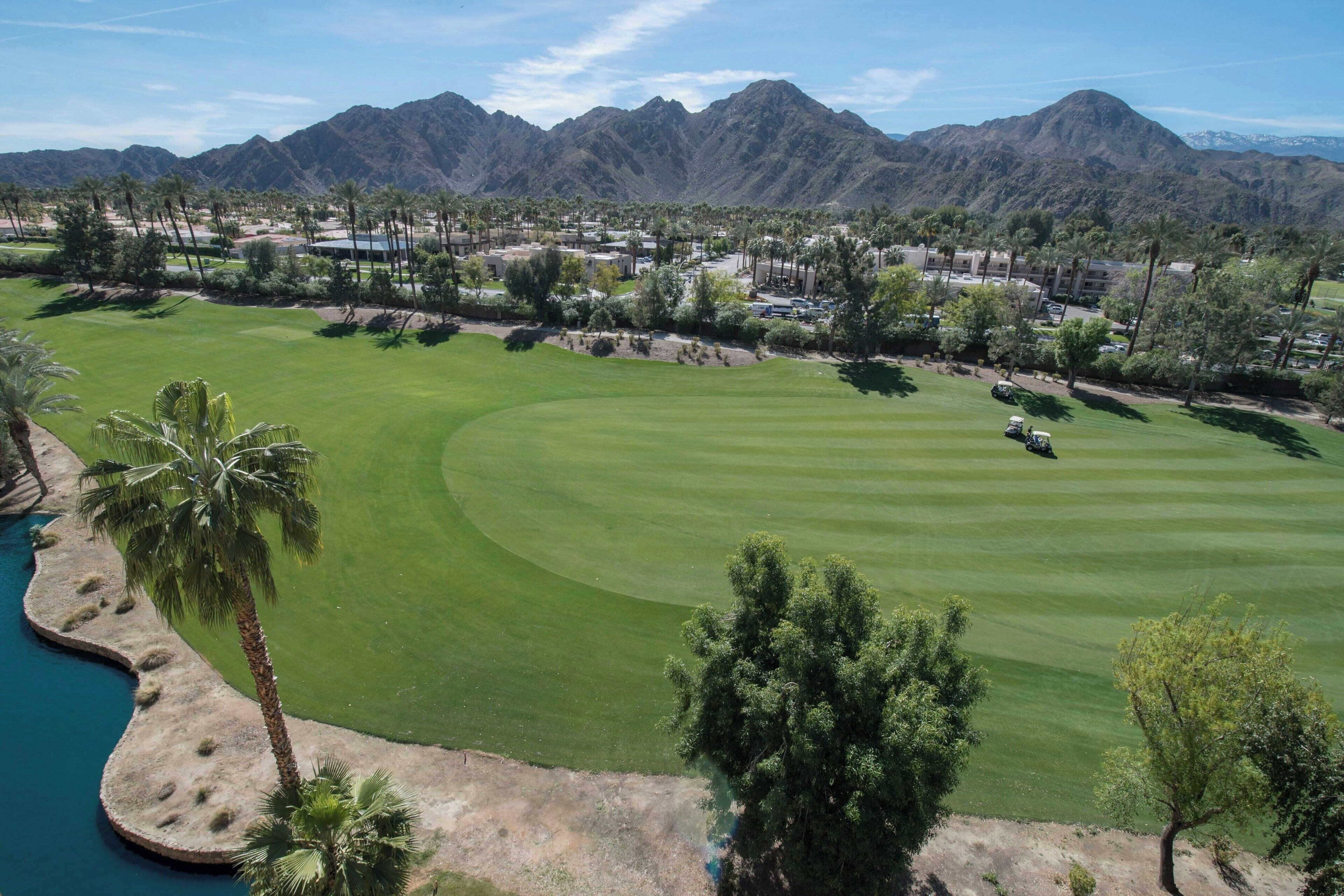 A golf course with a water feature, palm trees, and mountains in the background.