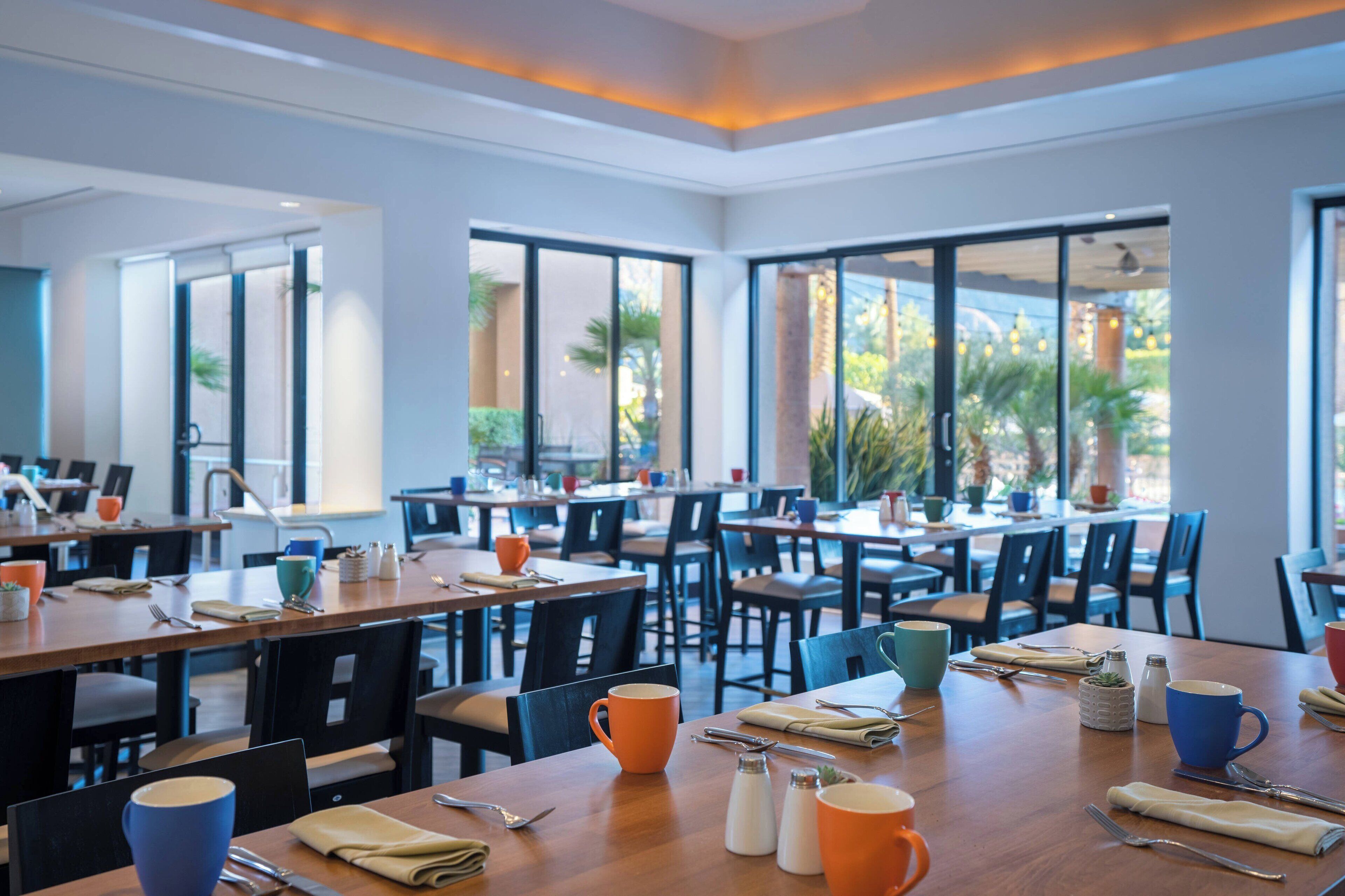 Un espace repas moderne doté de plusieurs tables et chaises, de grandes fenêtres et d’un intérieur lumineux.