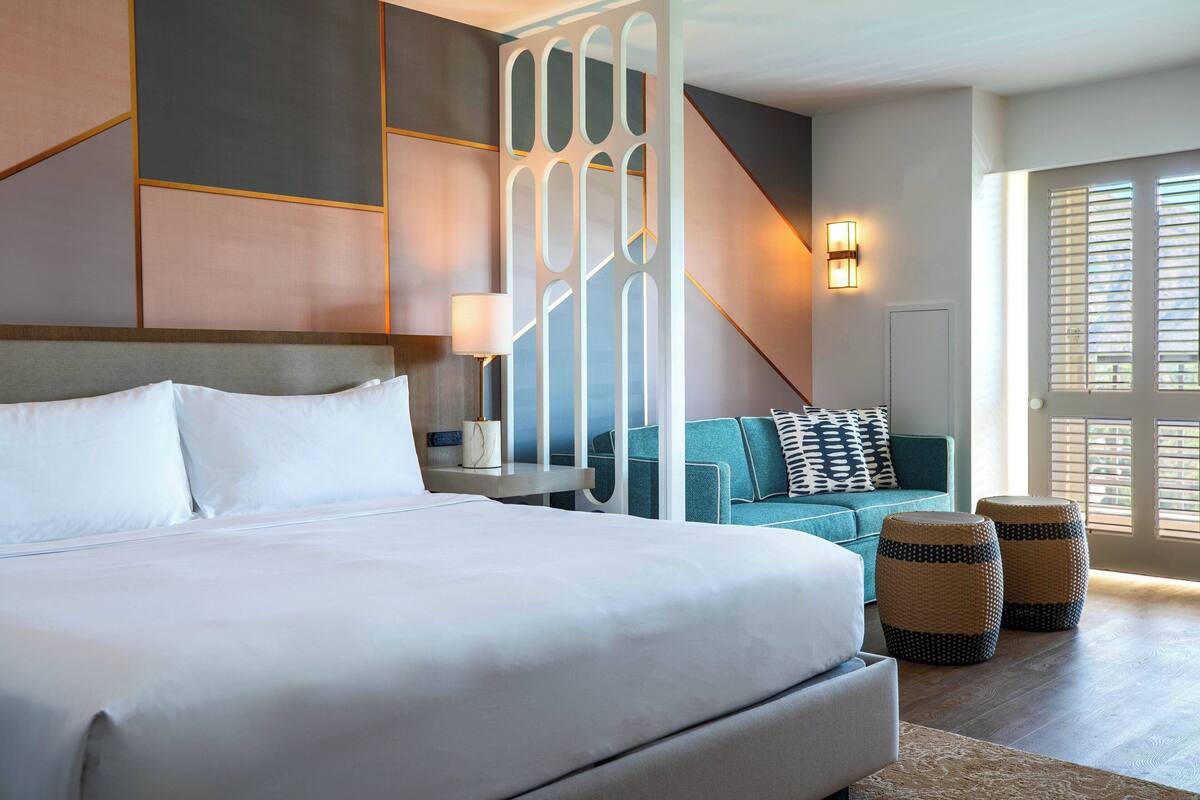 A modern hotel room with a large bed, a sofa, a decorative partition, and a view of the outside.