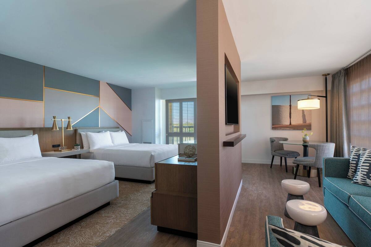 A modern hotel room with two beds, a sitting area with a sofa and chairs, a dining table, and a TV mounted on the wall.