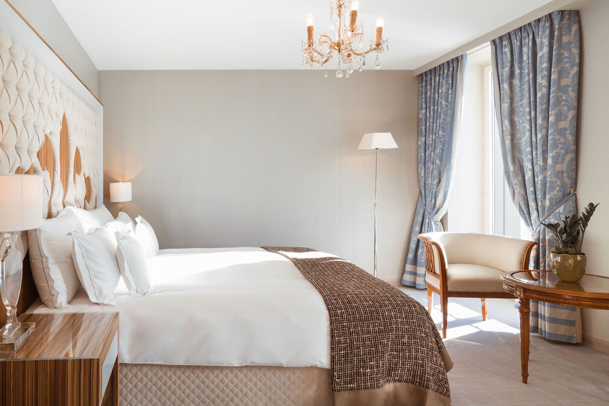 A hotel room with a large bed, a sitting area with a chair and a table, a chandelier, and a large window with curtains.