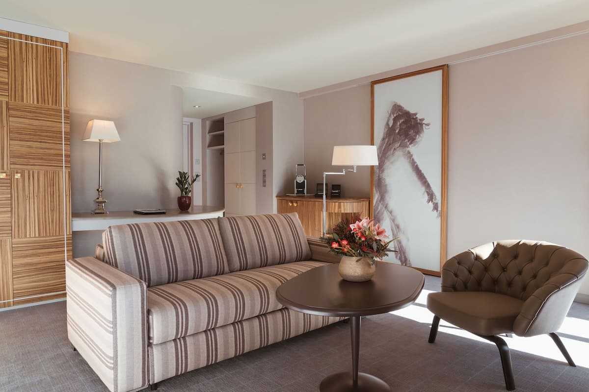 A modern living room with a striped sofa, a round table with a vase of flowers, a tufted armchair, and a large abstract painting on the wall.