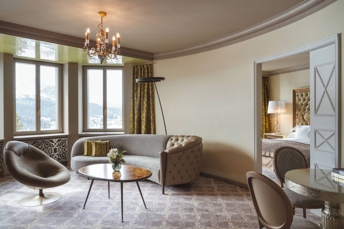 A modern hotel room with a curved sofa, a round coffee table, a tufted armchair, a dining table, and a chandelier.