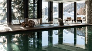 A modern indoor pool with clear glass walls, a stone wall, and a view of snowy mountains.