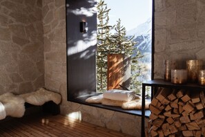 A cozy room with a stone wall, a bench, a window with a view of trees and mountains, and a stack of firewood.