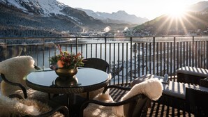 A balcony with a view of a lake and mountains, furnished with wicker chairs and a table.