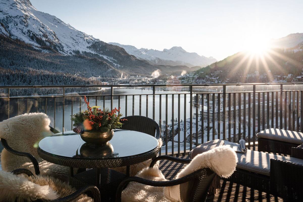 A balcony with a view of a lake and mountains, furnished with wicker chairs and a table.