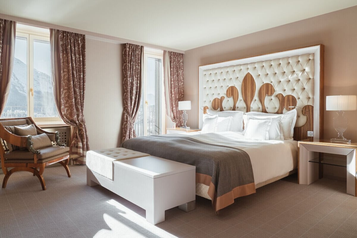 A spacious bedroom with a large bed, a sitting area with a chair and ottoman, and a view of the outdoors through large windows.