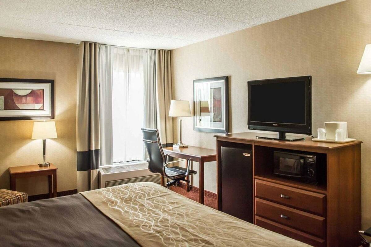 Habitación de hotel con cama, escritorio, silla, televisor, microondas y una mesita de noche con lámpara.