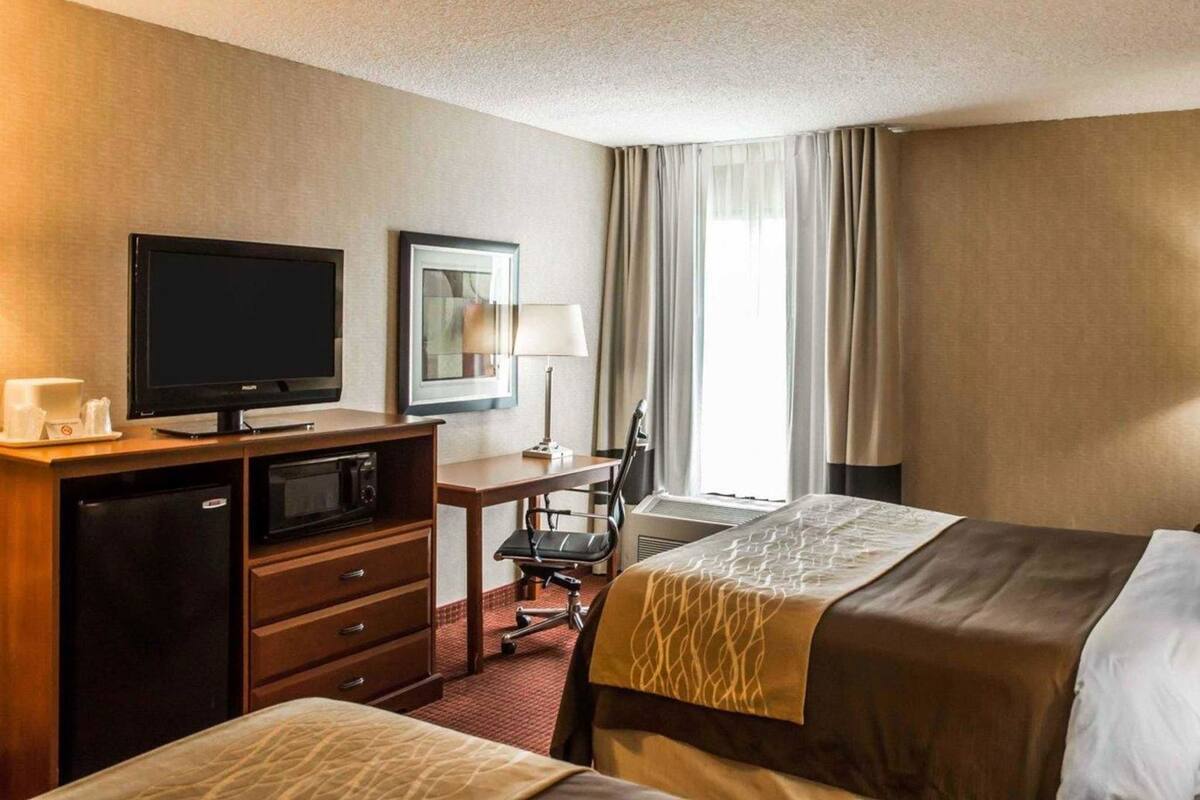 Una habitación de hotel con cama, escritorio con computadora, televisor, refrigerador, silla y ventana con cortinas.
