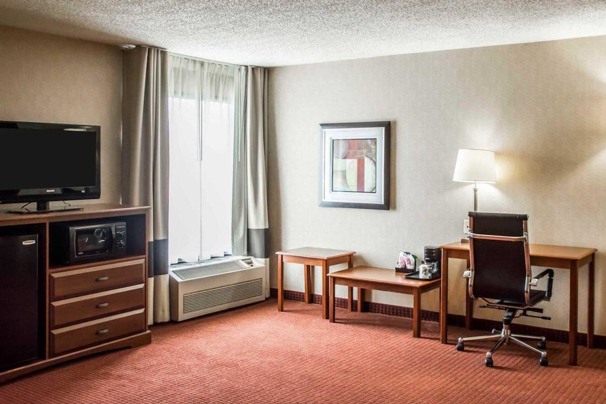 A hotel room with a television, air conditioning unit, desk, chair, and a framed picture on the wall.