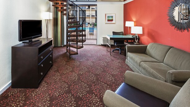 A compact living room with a flat-screen TV, a sofa, a desk with a chair, and a spiral staircase. - Holiday Inn Tyler-Conference Center by IHG (Tyler)