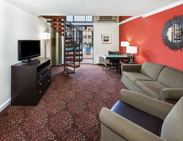 A compact living room with a flat-screen TV, a sofa, a desk with a chair, and a spiral staircase. - Holiday Inn Tyler-Conference Center by IHG (Tyler)
