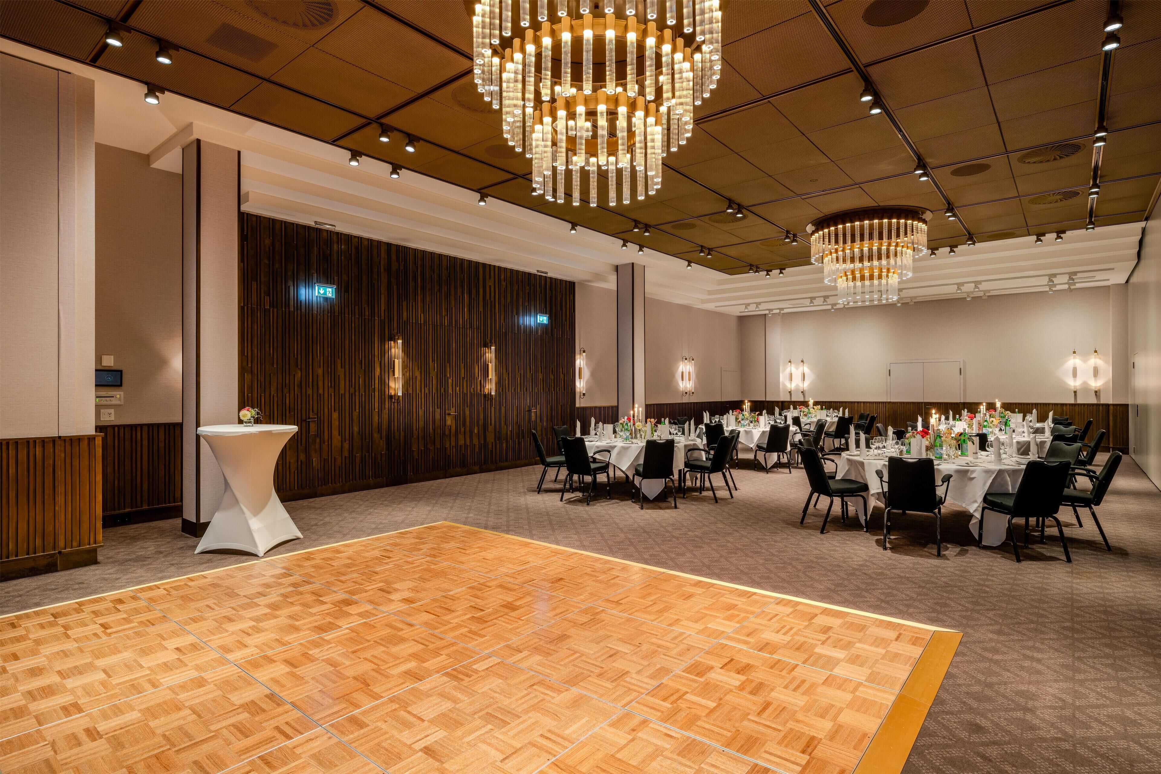 A spacious banquet hall with a polished wooden floor, multiple round tables set for dining, and elegant chandeliers.