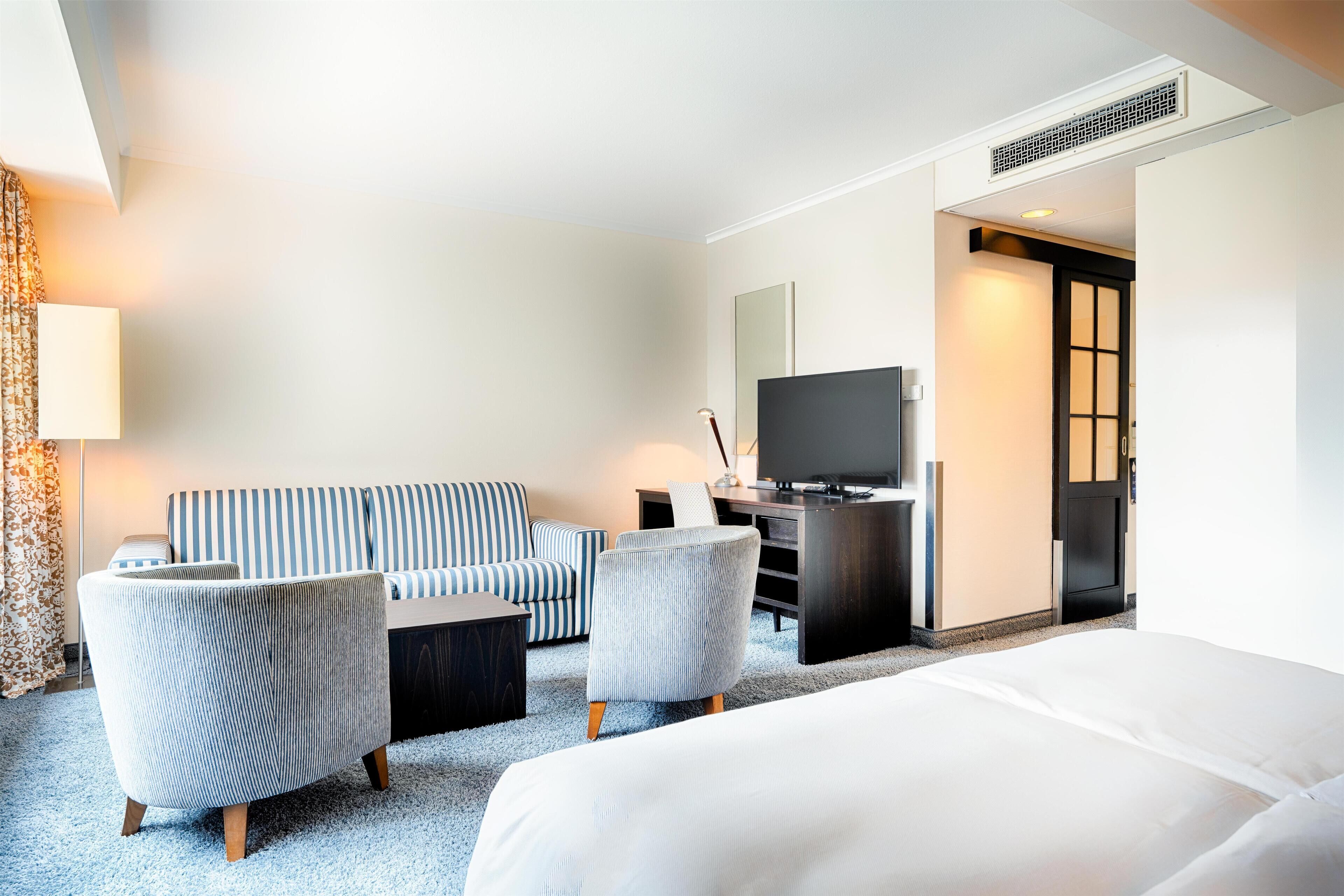 A hotel room with a bed, a sofa, two armchairs, a desk with a television, and a door leading to another room.