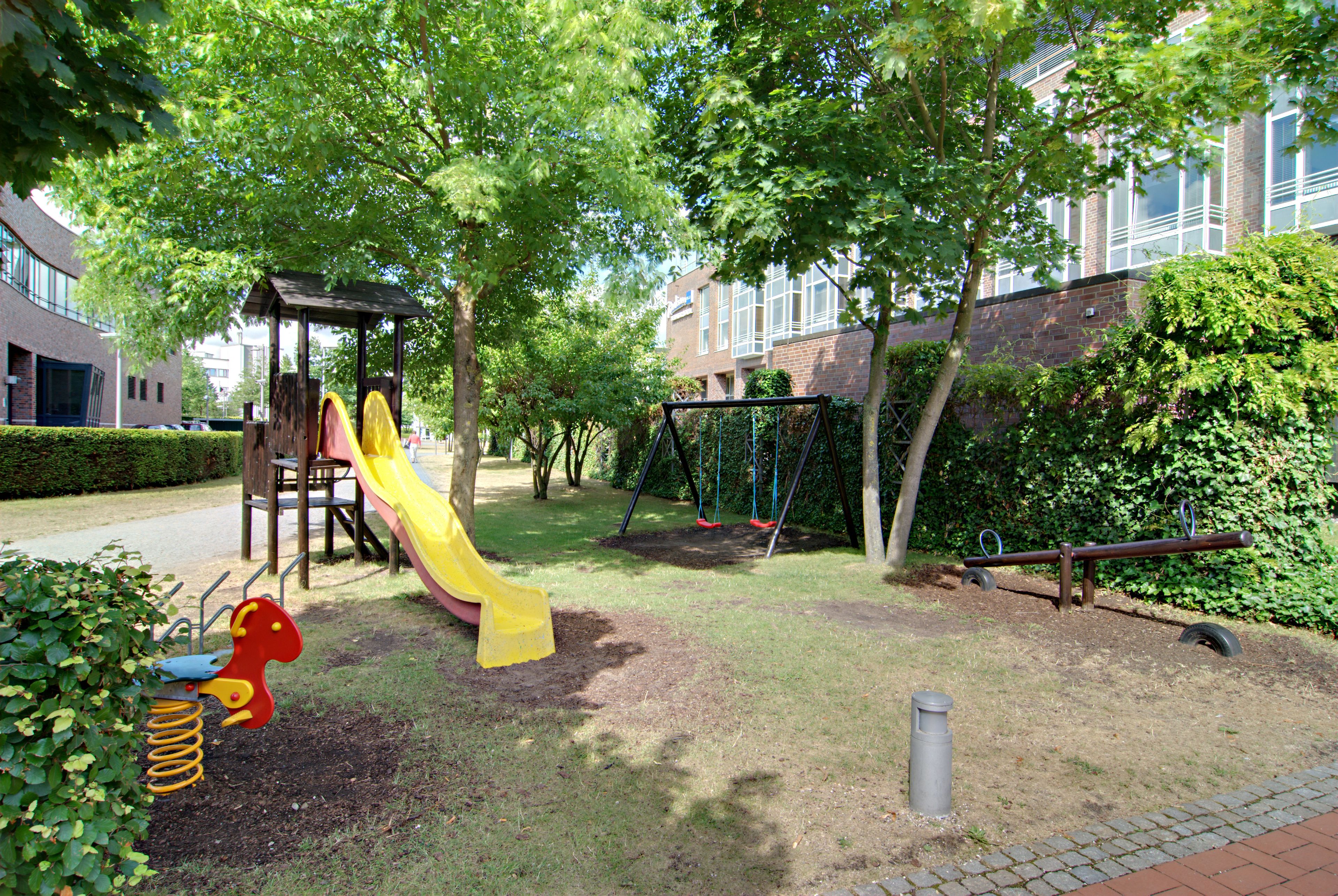A playground with a slide and swings, surrounded by trees and a building in the background.