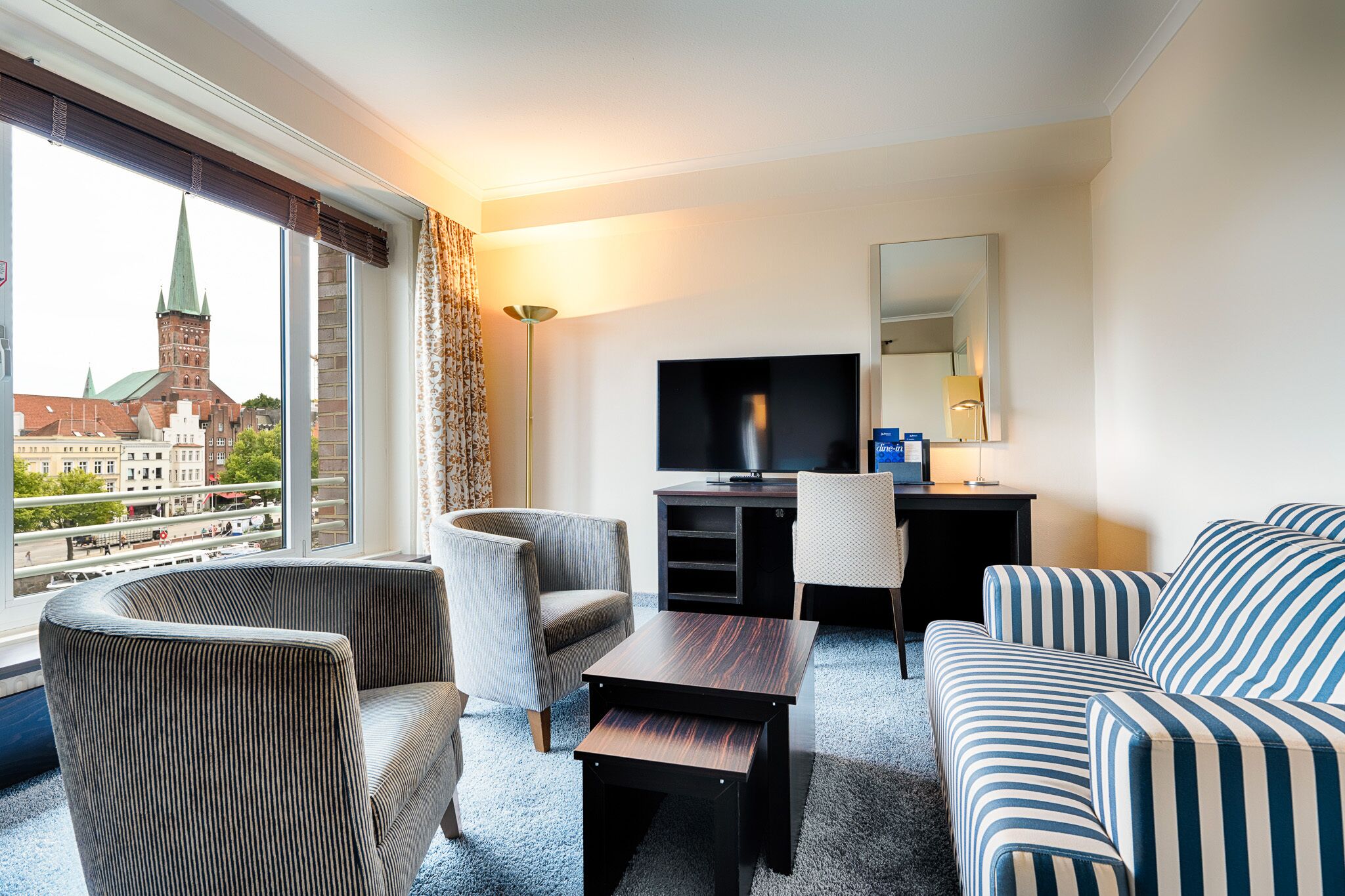 Habitación de hotel con un ventanal grande que ofrece vistas a un edificio histórico, un televisor de pantalla plana, un sofá, una silla y una mesita.
