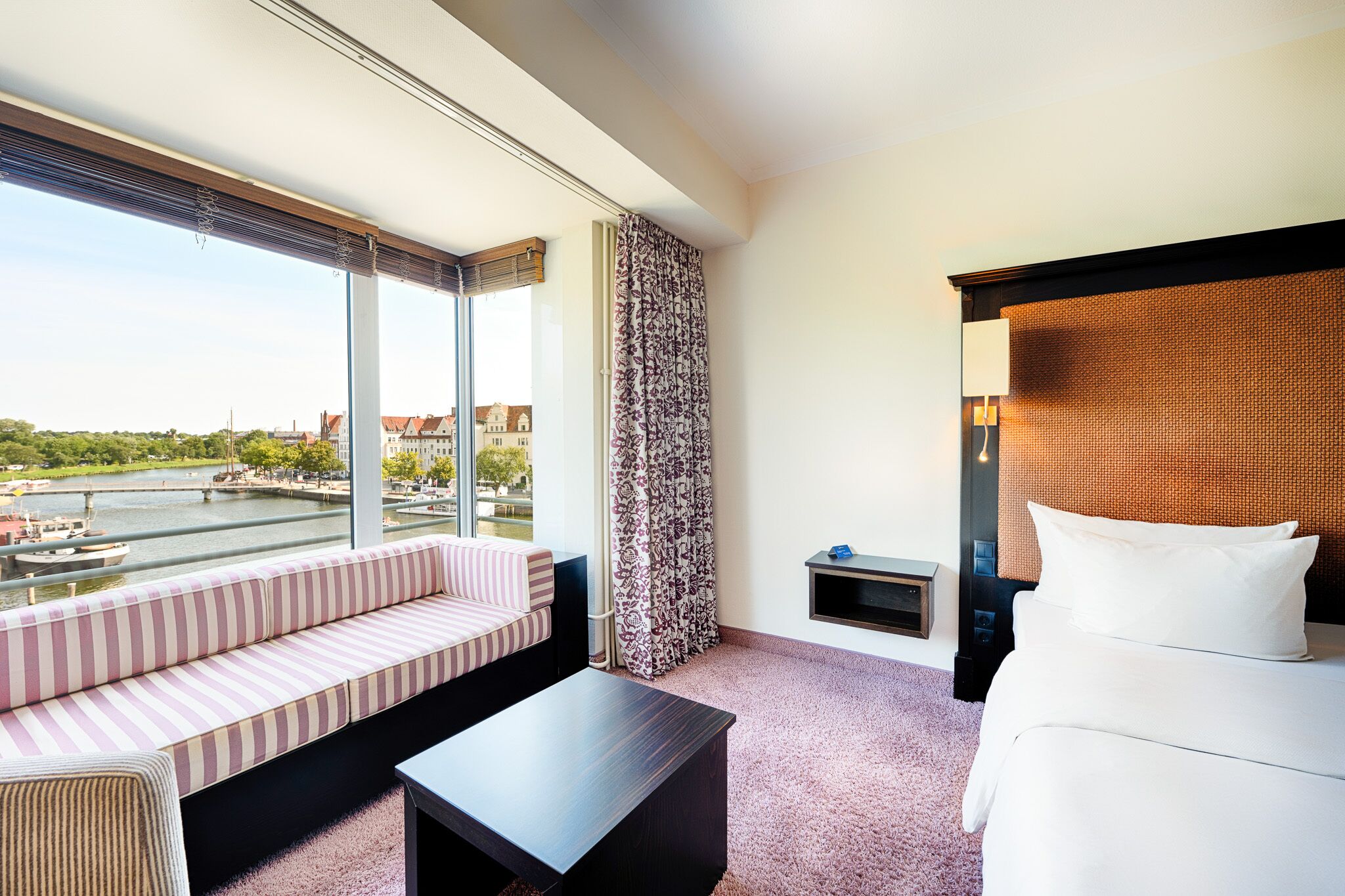 Habitación de hotel con un ventanal grande, cama, sofá, una mesita y una mesilla de noche.