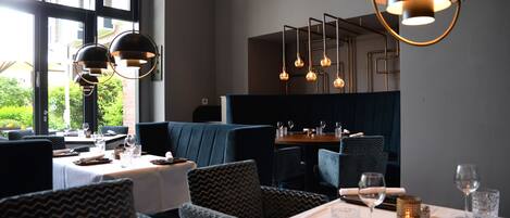 A restaurant with a modern interior, featuring dark blue upholstered seating, white tablecloths, and pendant lighting.