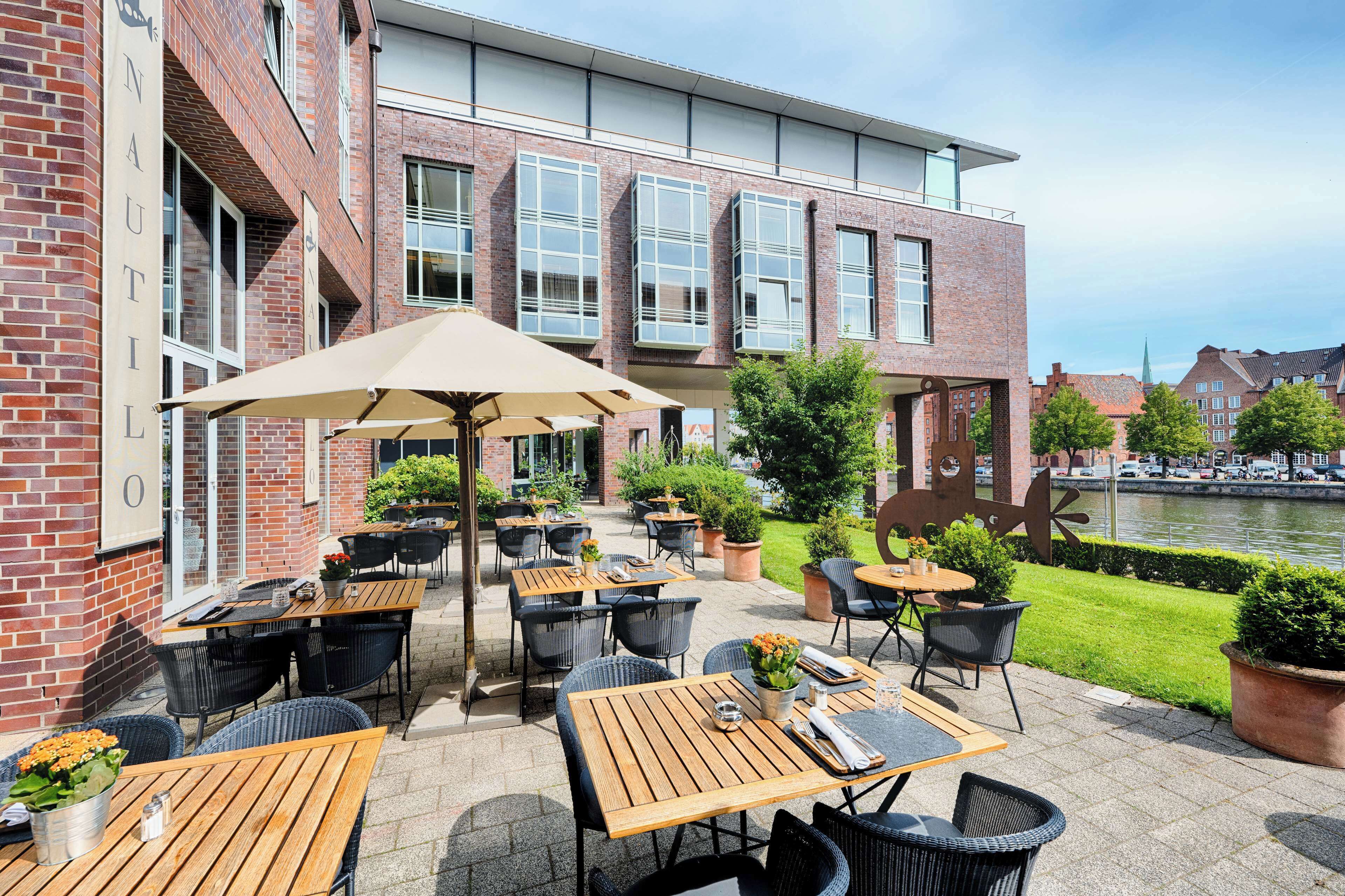 An outdoor patio with wooden tables and chairs, potted plants, and a view of a river.