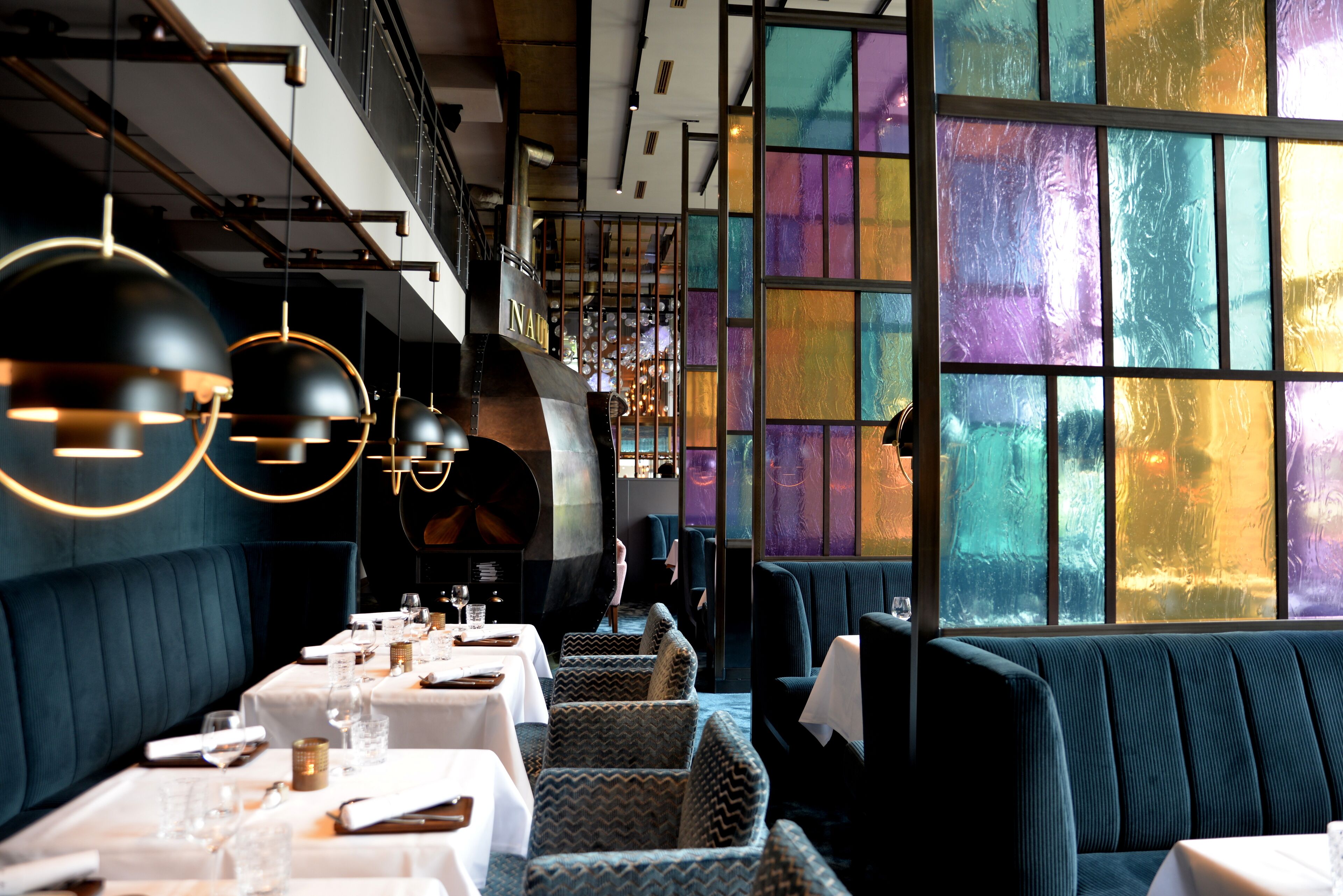 A restaurant with a modern interior, featuring a long dining area with tables and chairs, a high ceiling with exposed beams, and large windows with colorful stained glass.