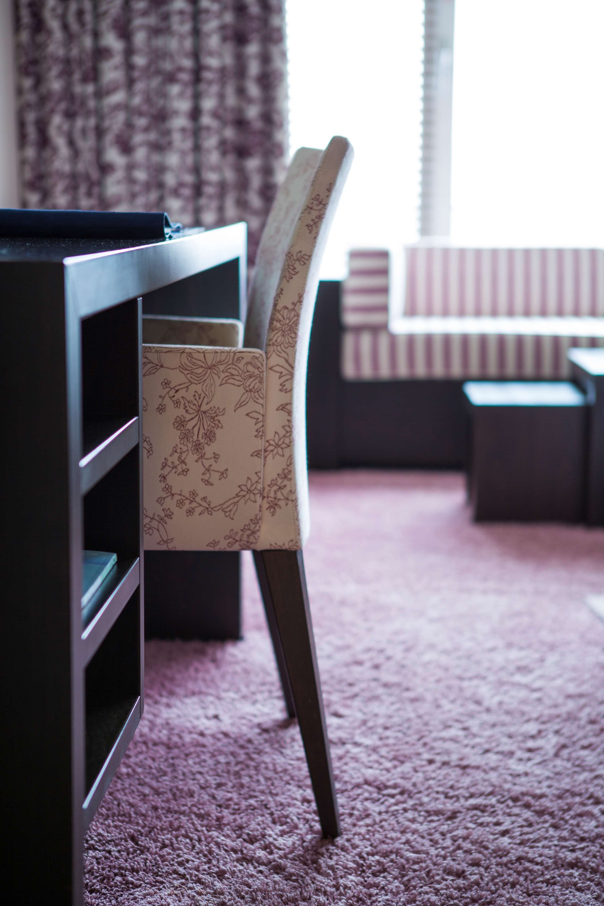 A room with a desk, a chair with floral upholstery, a striped couch, and a window with curtains.