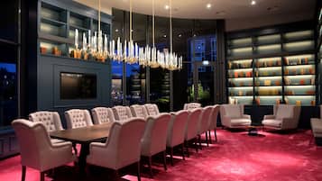 A modern dining area with a long table and upholstered chairs, a chandelier, and bookshelves with books.